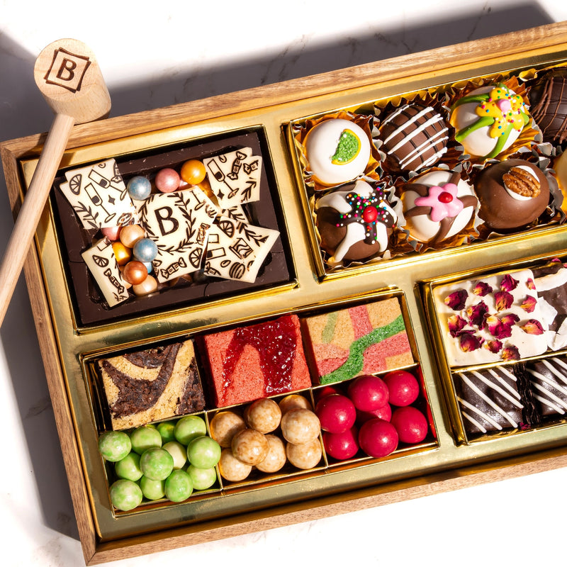 Wooden chocolate dessert board with white and dark chocolate pieces, decorated truffles, colorful candies, and fruit gummies arranged in gold compartments with a wooden smashing mallet.