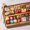 A gold-toned gift box filled with assorted holiday treats and chocolates, photographed from above on a light marble surface. The box contains decorated candies, candy canes, nuts, wrapped chocolates, and a small wooden mallet with a letter B.