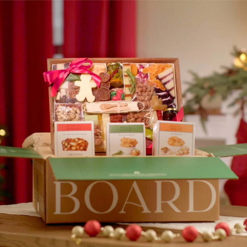 Holiday-themed cheese and charcuterie board with festive cheese shapes, nuts, dried fruits, and crackers, tied with a red ribbon and boxed with artisan crisps.