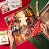 Hands holding a Boarderie charcuterie box featuring an assortment of cheeses, nuts, dried fruits, & fig spread, wrapped in branded ribbon