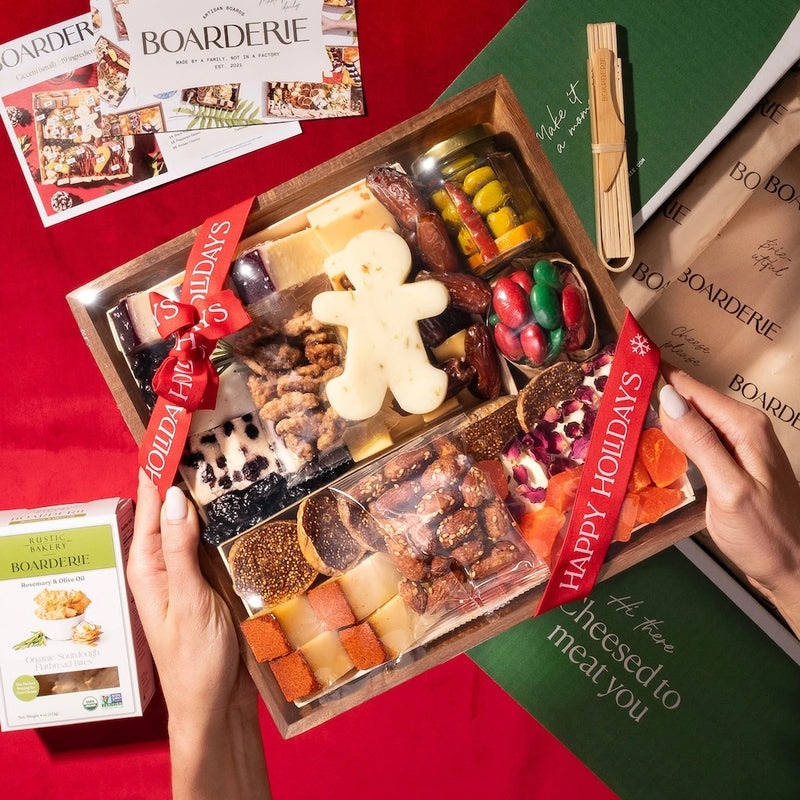 Hands holding a Boarderie charcuterie box featuring an assortment of cheeses, nuts, dried fruits, & fig spread, wrapped in branded ribbon