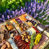 Close-up of a gourmet charcuterie board with meats, cheeses, nuts, and dried fruits in a garden setting with purple flowers.