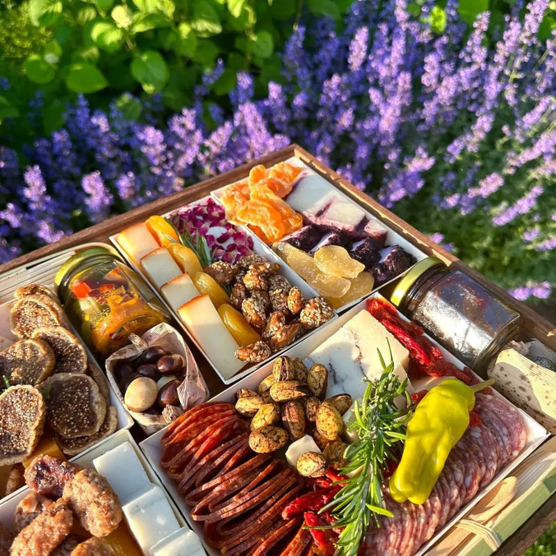 Close-up of a gourmet charcuterie board with meats, cheeses, nuts, and dried fruits in a garden setting with purple flowers.