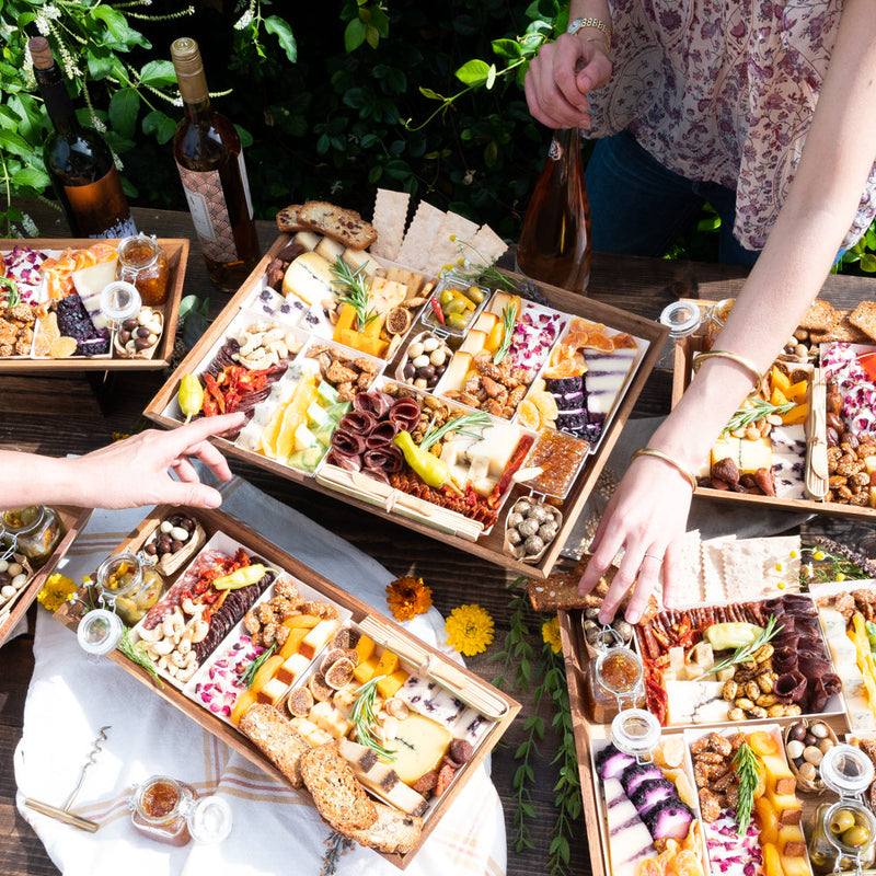 lively outdoor gathering showcasing an array of gourmet Boarderie charcuterie boards filled with artisan cheeses, nuts, dried fruits, & gourmet accompaniments, paired with bottles of wine