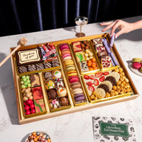 A top-down view of the Thinking of You Dessert Board Large, featuring a reusable acacia wood board filled with over 30 artisan sweets, including macarons, decorated truffles, and gourmet candies in elegant gold compartments. A hand holds a glass nearby, and a small wooden mallet rests next to a central chocolate box engraved with 