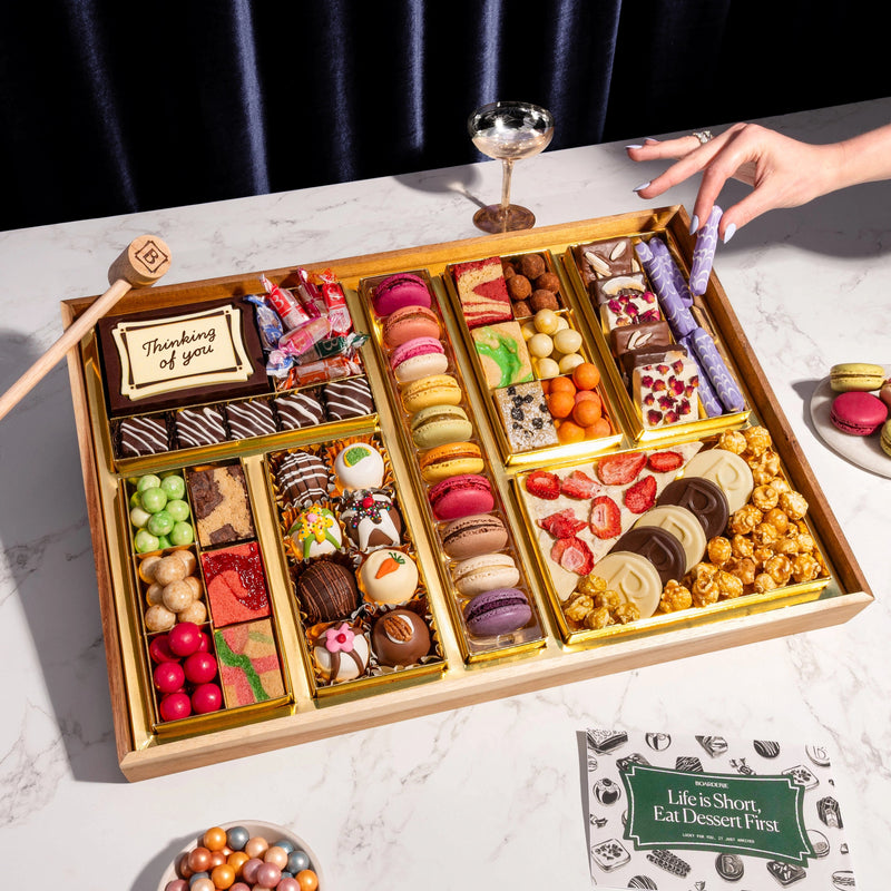 A top-down view of the Thinking of You Dessert Board Large, featuring a reusable acacia wood board filled with over 30 artisan sweets, including macarons, decorated truffles, and gourmet candies in elegant gold compartments. A hand holds a glass nearby, and a small wooden mallet rests next to a central chocolate box engraved with "Thinking of You."