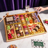 Large chocolate dessert board with macarons, decorated truffles, candies, strawberries, and popcorn arranged in gold compartments with wooden mallet and serving utensil.