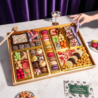 Large chocolate dessert board with macarons, decorated truffles, candies, strawberries, and popcorn arranged in gold compartments with wooden mallet and serving utensil.