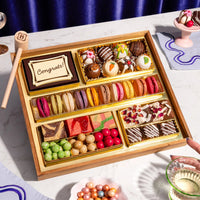 Assorted chocolates in a wooden box on a marble surface with a hand holding a glass.