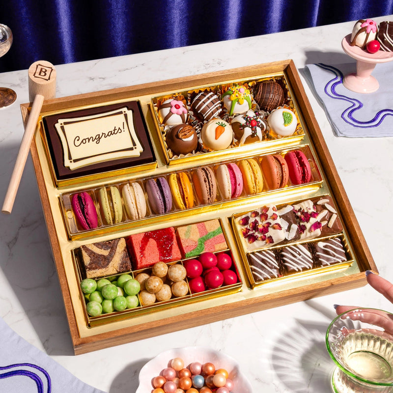 Assorted chocolates in a wooden box on a marble surface with a hand holding a glass.