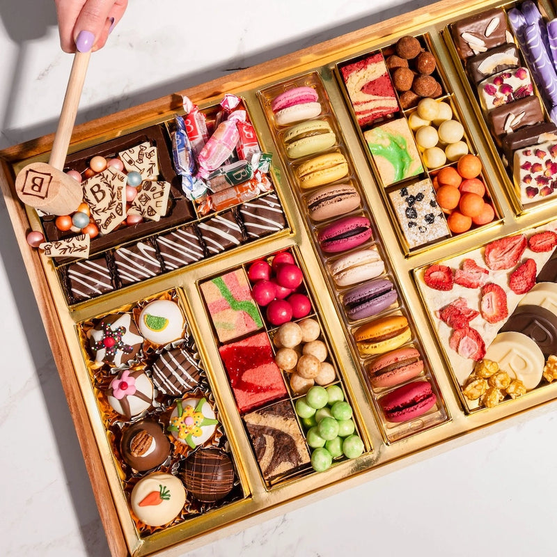 Valentines Day dessert board with wooden mallet, featuring macarons, decorated truffles, candies, strawberries, and gourmet treats in gold compartments for smashing.