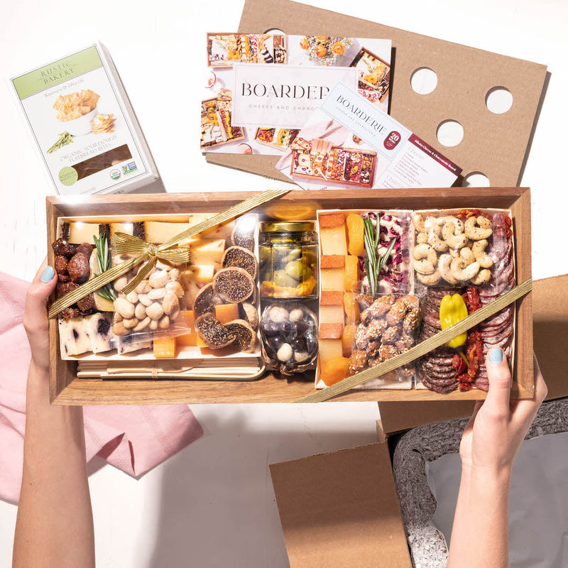 Hands holding a gift charcuterie board with cheese, figs, nuts, and crackers, packaged with Boarderie branding.