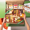 A charcuterie board gift box held by two hands over a green Boarderie Box, featuring an assortment of cured meats, cheeses, crackers, nuts, and fruits. The Boarderie Box is visible in the background.