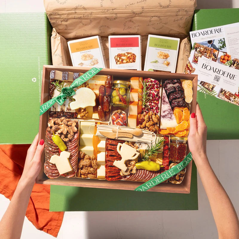 A charcuterie board gift box held by two hands over a green Boarderie Box, featuring an assortment of cured meats, cheeses, crackers, nuts, and fruits. The Boarderie Box is visible in the background.
