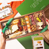 A wooden charcuterie board filled with cheeses, cured meats, nuts, olives, and crackers, held by two hands with a green 'The Cheese Meat You' gift box and a Boarderie box visible in the background.