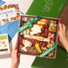 A wooden charcuterie board gift box held by two hands, featuring cured meats, cheeses, crackers, olives, and garnishes, surrounded by green packaging and printed cards.