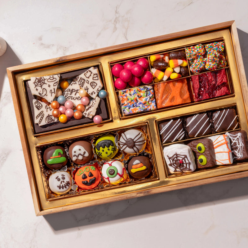 A wooden gift box filled with assorted Halloween-themed chocolates, candies, and treats, photographed from above on a light marble surface.