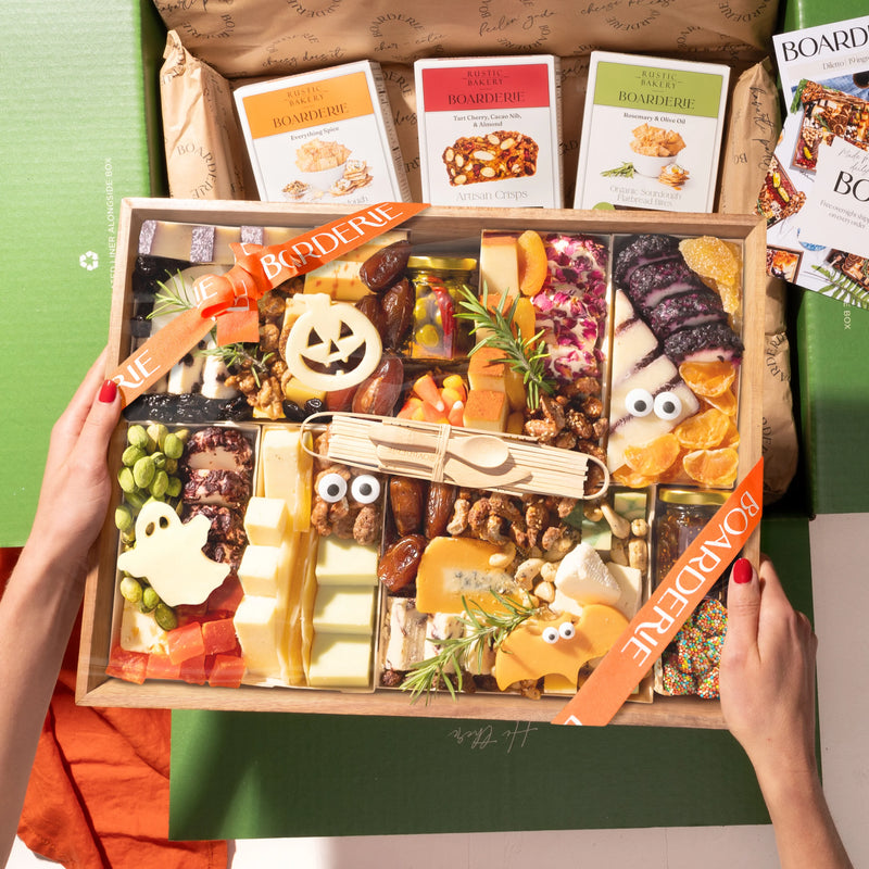 A hands-on view of the Boarderie Large Halloween All Cheese ' gift box being held. The wooden tray contains an assortment of cheeses, crackers, candy corn.