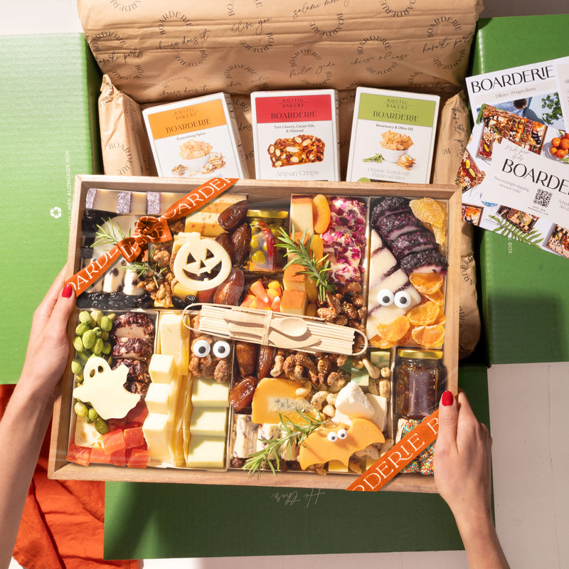 A hands-on view of the Boarderie Large Halloween All Cheese ' gift box being held. The wooden tray contains an assortment of cheeses, crackers, candy corn.
