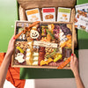 A hands-on view of the Boarderie Large Halloween Cheese & CharBOOterie' gift box being held. The wooden tray contains an assortment of cheeses, cured meats, crackers, candy corn.