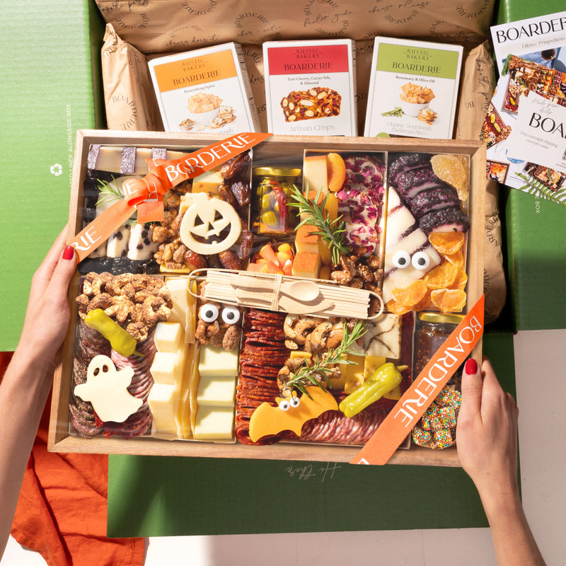 A hands-on view of the Boarderie Large Halloween Cheese & CharBOOterie' gift box being held. The wooden tray contains an assortment of cheeses, cured meats, crackers, candy corn.