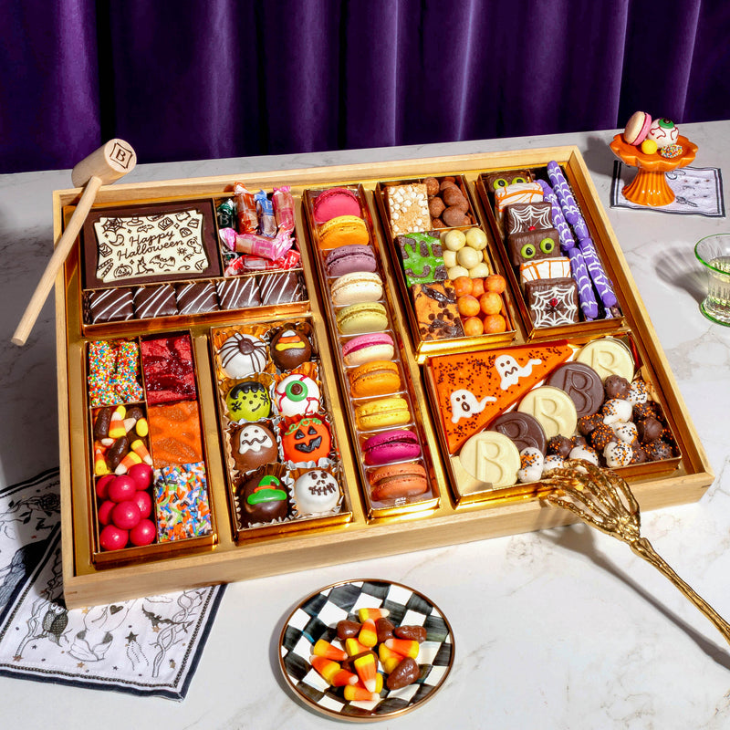 A large wooden dessert display filled with a fully-arranged assorted Halloween-themed chocolates, macarons, truffles, candies, chocolate covered oreos and treats, styled on a white marble surface with a purple curtain background. Includes a chocolate smash box filled with candies.