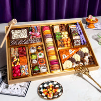 Large Halloween dessert board loaded with macarons, chocolate-covered pretzels, cake pops, monogrammed chocolate coins, a ghost pizza slice cookie, and hand-painted spooky truffles, with a golden skeleton hand reaching in.