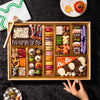 Overhead shot of a large Halloween dessert board on black backdrop with a hand picking up a truffle, showing mummy brownie bites, spiderweb cake bites, purple bone-shaped candy, macarons, and a ghost pizza slice cookie.