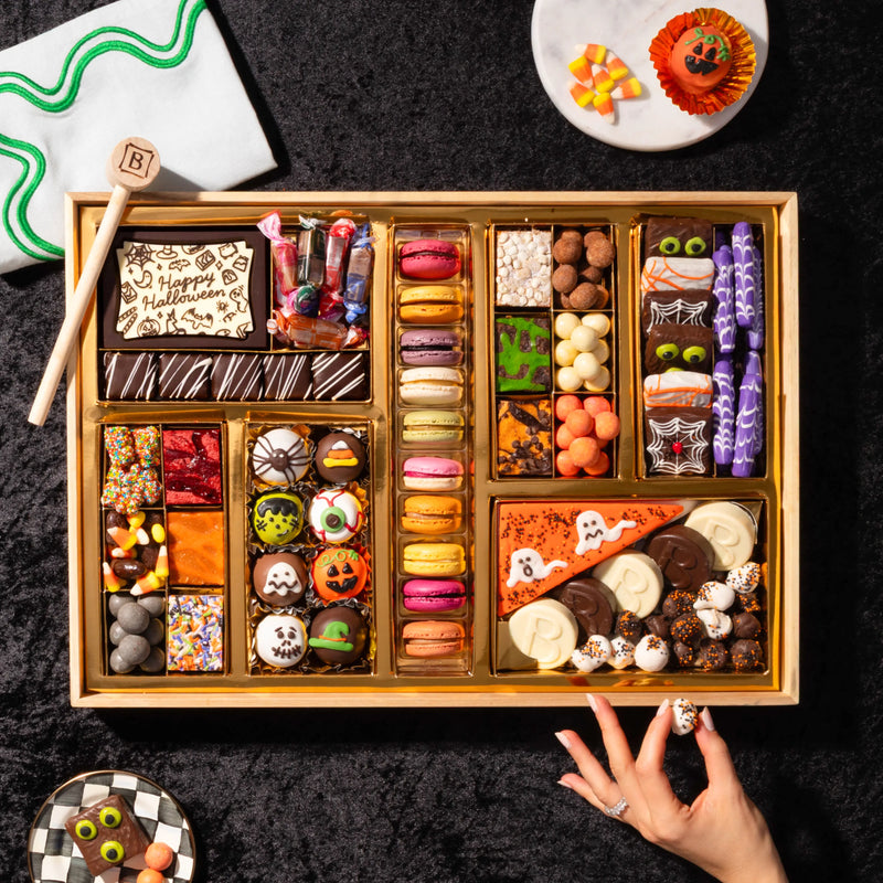 Overhead shot of a large Halloween dessert board on black backdrop with a hand picking up a truffle, showing mummy brownie bites, spiderweb cake bites, purple bone-shaped candy, macarons, and a ghost pizza slice cookie.