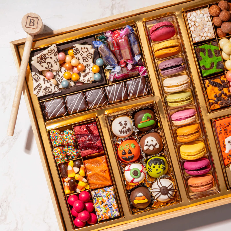 Close-up angled view of a large Halloween dessert board with Happy Halloween chocolate bark, wrapped hard candies, mummy brownie bites, an assorted macaron column, and hand-decorated skull, pumpkin, and eyeball truffles.