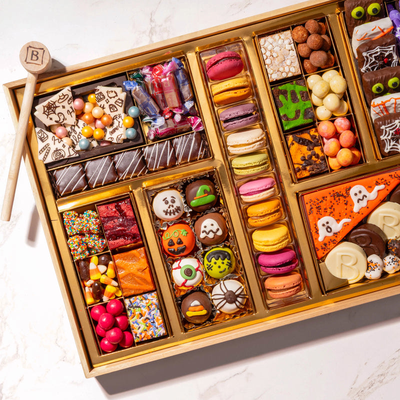 Close-up angled view of a large Halloween dessert board with Happy Halloween chocolate bark, wrapped hard candies, mummy brownie bites, an assorted macaron column, and hand-decorated skull, pumpkin, and eyeball truffles.