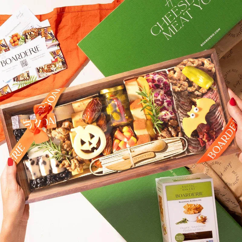 A hands-on view of the Boarderie Medium Halloween All Cheese' gift box being held. The wooden tray contains an assortment of cheeses, crackers, candy corn.