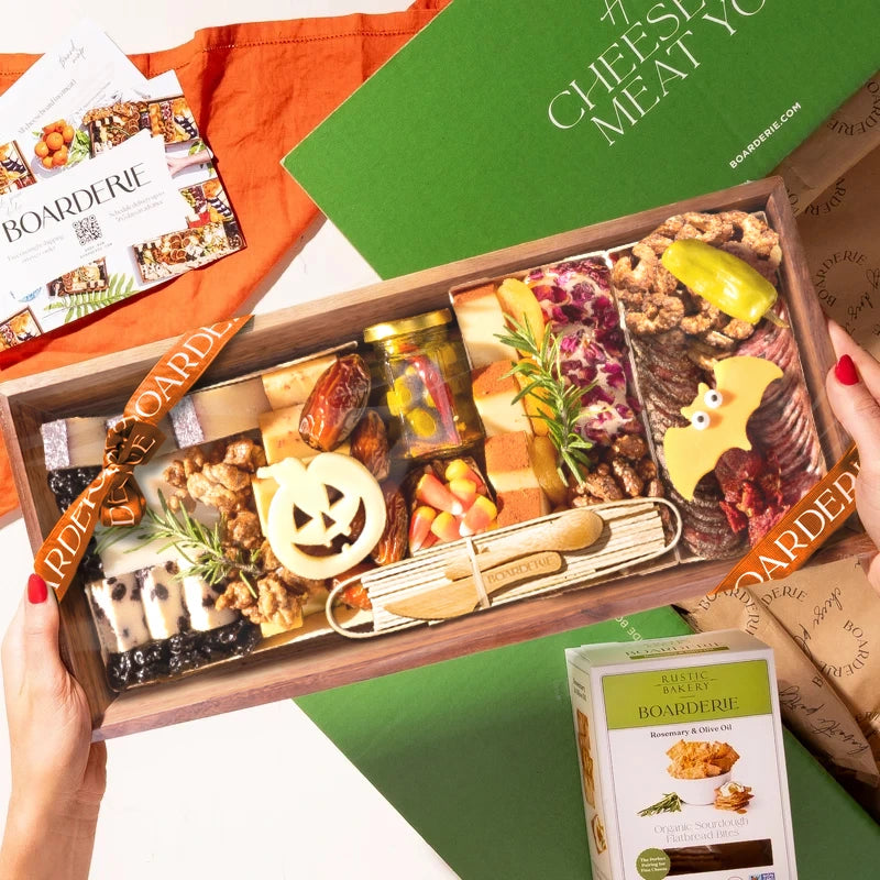A hands-on view of the Boarderie Medium Halloween Cheese & CharBOOterie' gift box being held. The wooden tray contains an assortment of cheeses, cured meats, crackers, candy corn.