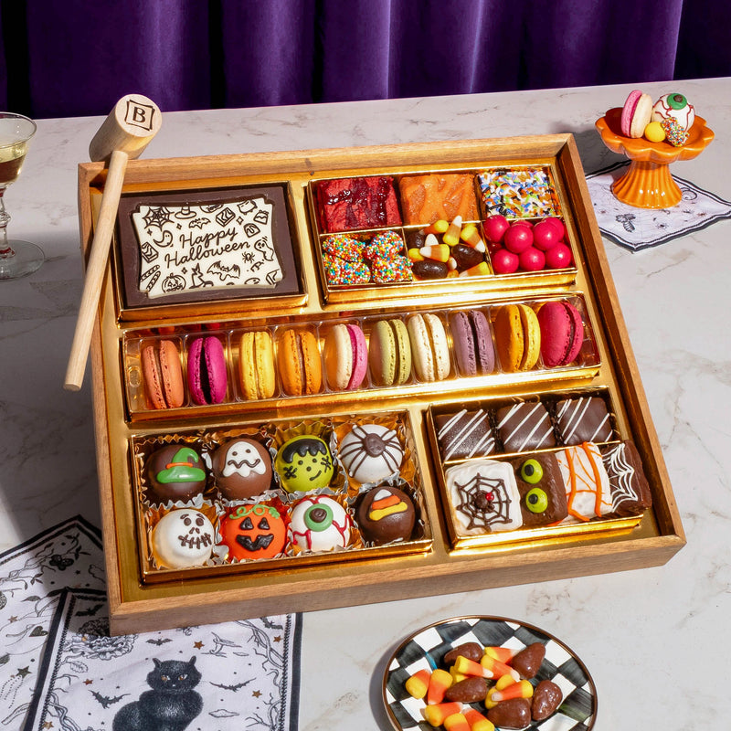 A wooden gift box filled with assorted Halloween-themed treats, including macarons, halloween themed truffles, decorated chocolate candies, candy corn, spiderweb toffee, and a chocolate smash box that says 'Happy Halloween.' The box is styled on a marble surface with purple curtains in the background.