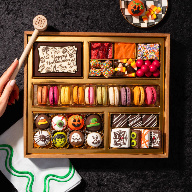 Overhead view of a medium Halloween dessert board on a dark backdrop, with a hand reaching in, showing hand-decorated truffles, mummy-striped brownie bites, and a rainbow row of French macarons.