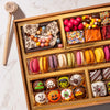 Close-up of a medium Halloween dessert board showing a shattered "Happy Halloween" chocolate bark with metallic candy pearls, gumballs, candy corn, and a row of French macarons alongside hand-painted spooky truffles.