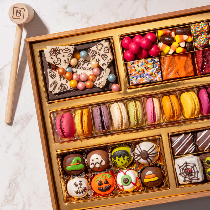 Close-up of a medium Halloween dessert board showing a shattered "Happy Halloween" chocolate bark with metallic candy pearls, gumballs, candy corn, and a row of French macarons alongside hand-painted spooky truffles.