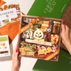 A hands-on view of the Boarderie Classic Halloween All Cheese' gift box being held. The wooden tray contains an assortment of cheeses, cured meats, crackers, candy corn.