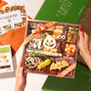 A hands-on view of the Boarderie Classic Cheese & Charcuterie' gift box being held. The wooden tray contains an assortment of cheeses, cured meats, crackers, candy corn.