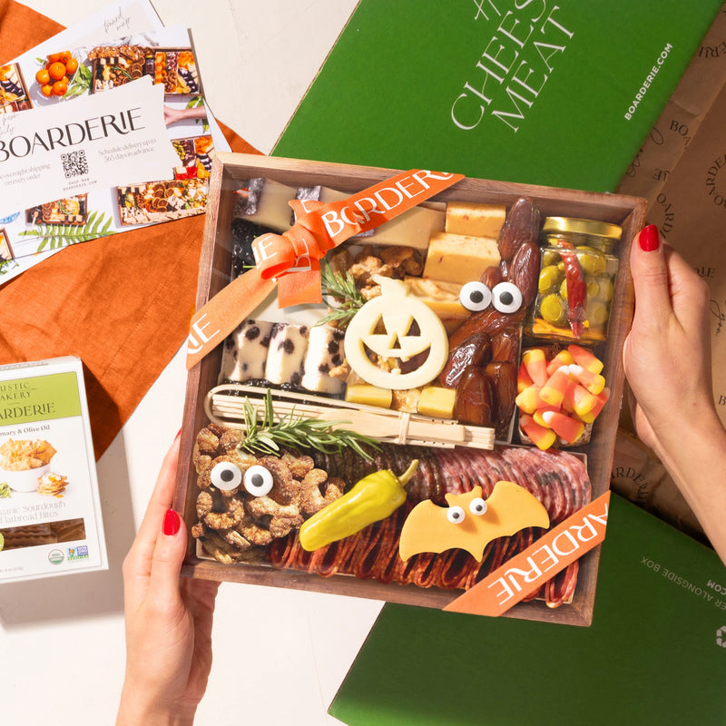 A hands-on view of the Boarderie Classic Cheese & Charcuterie' gift box being held. The wooden tray contains an assortment of cheeses, cured meats, crackers, candy corn.