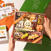 A hands-on view of the Boarderie Classic Cheese & Charcuterie' gift box being held. The wooden tray contains an assortment of cheeses, cured meats, crackers, candy corn.