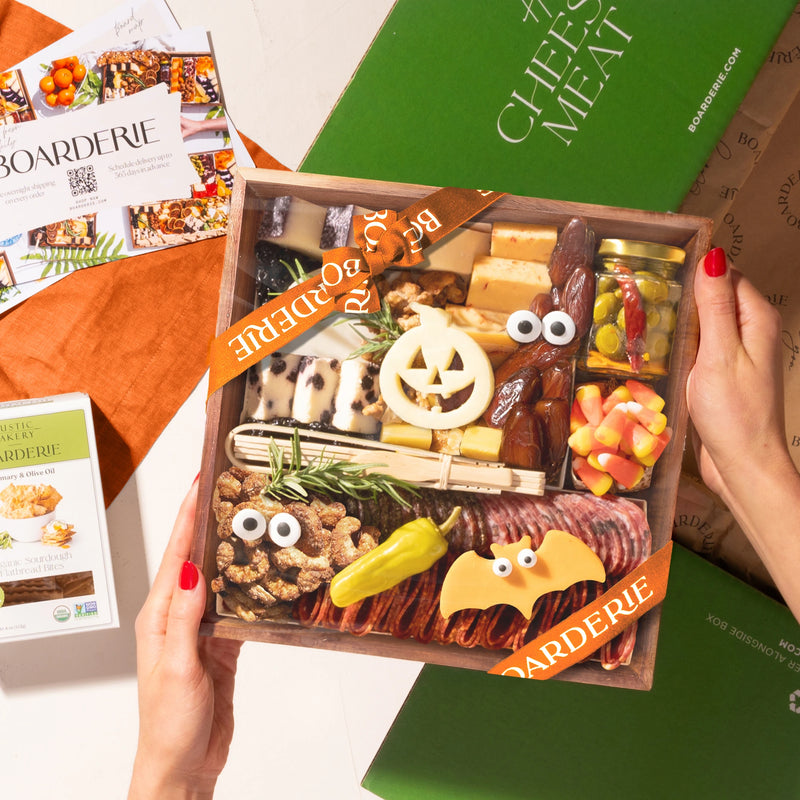 A hands-on view of the Boarderie Classic Cheese & Charcuterie' gift box being held. The wooden tray contains an assortment of cheeses, cured meats, crackers, candy corn.