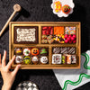 Small Halloween dessert board on a dark backdrop with a hand reaching for hand-painted spider and Frankenstein truffles, alongside brownie bites, candy corn, and a "Happy Halloween" chocolate plaque in a gold-trimmed wooden tray.