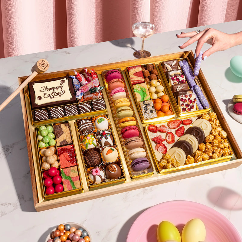 Large Easter dessert board filled with macarons, chocolate truffles, candies, caramel popcorn, and a "Happy Easter" chocolate plaque, styled on a marble surface with pastel accents