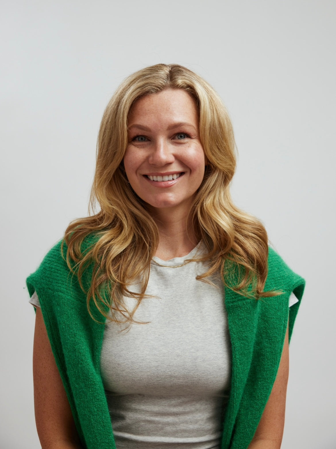 Kassy Edwards with long blonde hair smiling, wearing a gray shirt and green sweater draped over her shoulders against a neutral background.