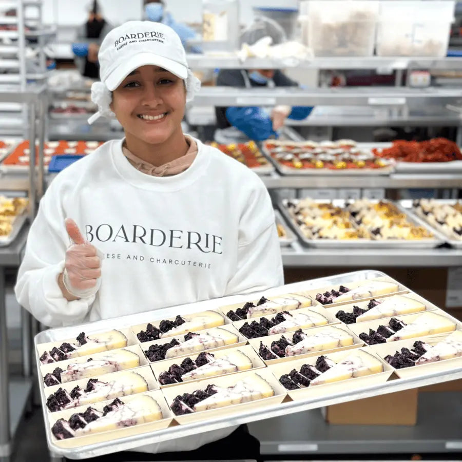 Charcuterie worker presenting a tray of cheese slices topped with blueberries in a food preparation facility.