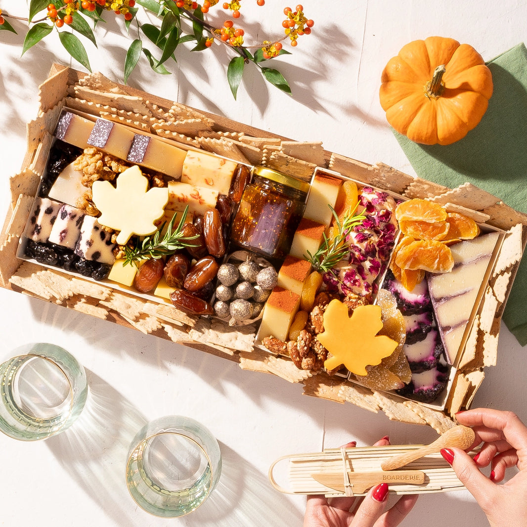 Autumn-themed charcuterie board with assorted cheeses, dried fruit, nuts, crackers, & a maple leaf-shaped cheese, decorated with fall accents