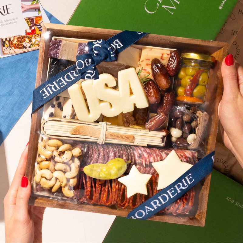 A Labor Day USA charcuterie board gift held by two hands, featuring an assortment of cheeses, meats, and nuts. 