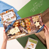 An overhead view of a Labor Day USA charcuterie board gift box being held by two hands, placed next to an open green  labeled. The board is filled with various cheeses, crackers, nuts, and fruits.