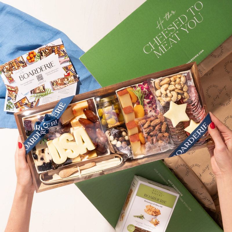 An overhead view of a Labor Day USA charcuterie board gift box being held by two hands, placed next to an open green  labeled. The board is filled with various cheeses, crackers, nuts, and fruits.